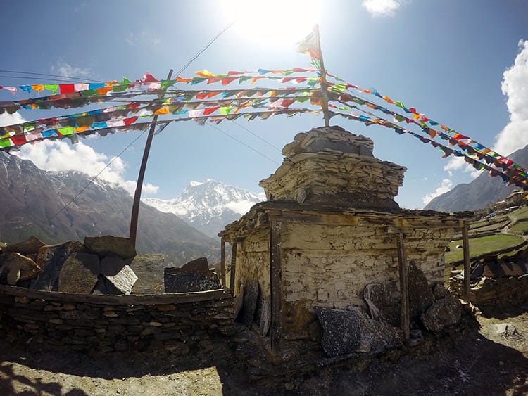 A small stone stupa stands with colorful prayer flags blowing in the wind and the Annapurna range in the background