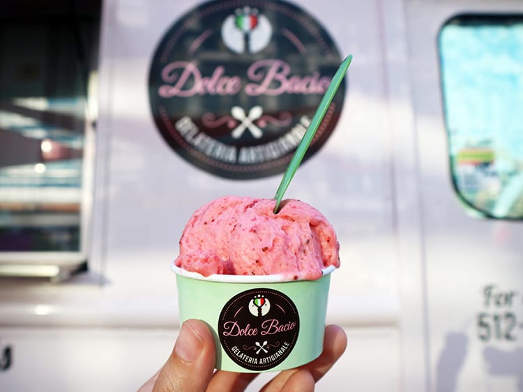 A cup of strawberry gelato held up in front of the Dolce Bacio food truck in Austin