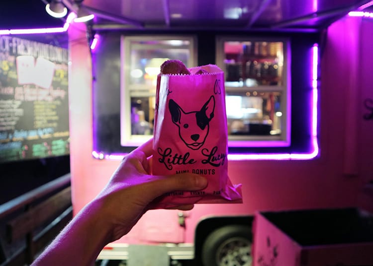 A bag of donuts held up in front of the pink Little Lucy's food truck at night