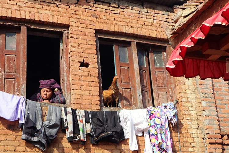 A woman and her chickens look out the window and watch people walking by below in Panauti