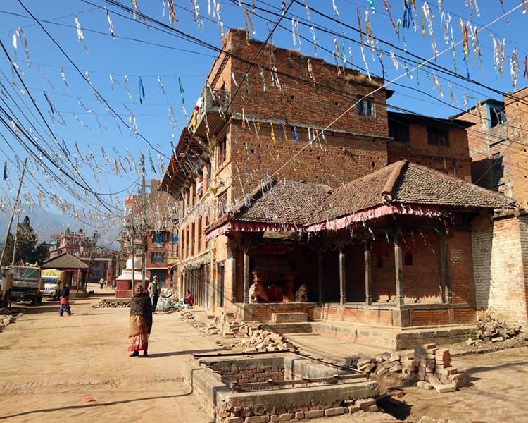 An old woman walks down the street while silver garland hangs over the street and blows in the wind in Panauti