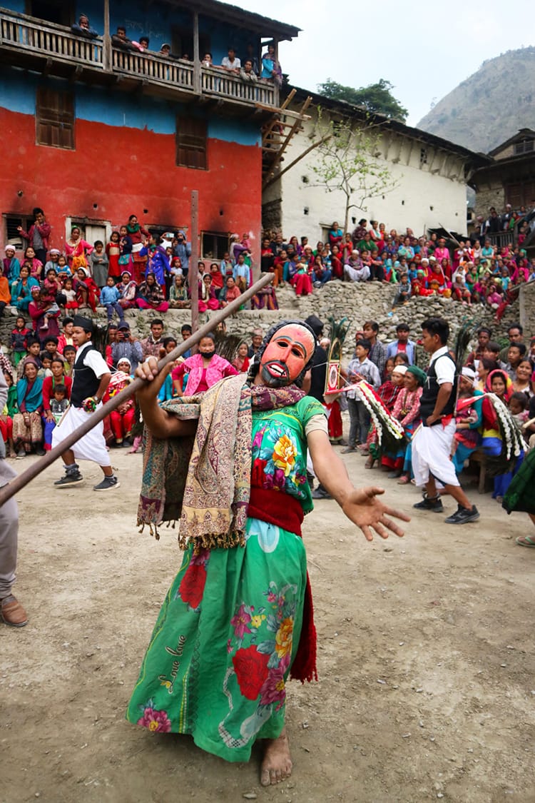 A man wearing a green dress and a bright orange mask does a traditional stick dance