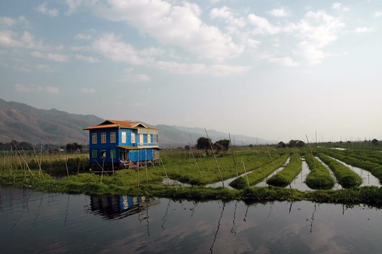 A blue house sits in the floating gardens in Michelle Della Giovanna from Full Time Explorer sits in a blue boat on Inle Lake, Myanmar