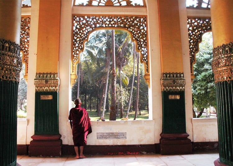 A Burmese monk in maroon robes stands in the elaborate doorway of a temple in Yangon, Myanmar
