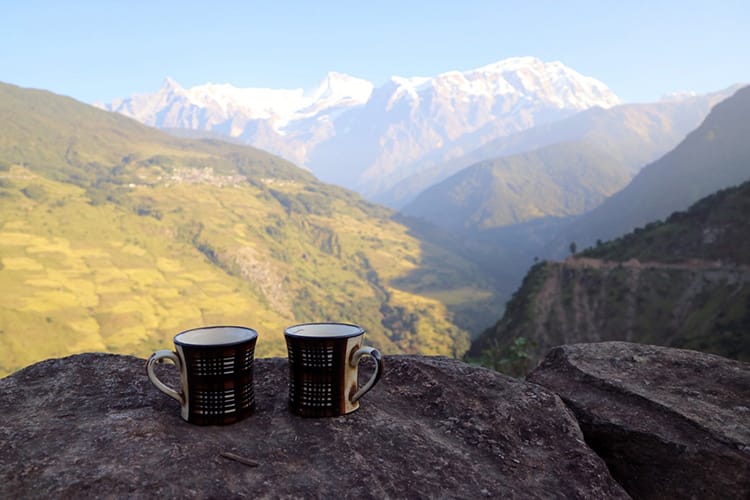Two cups of tea sit on a stone wall with a view of the Annapurna range in the background
