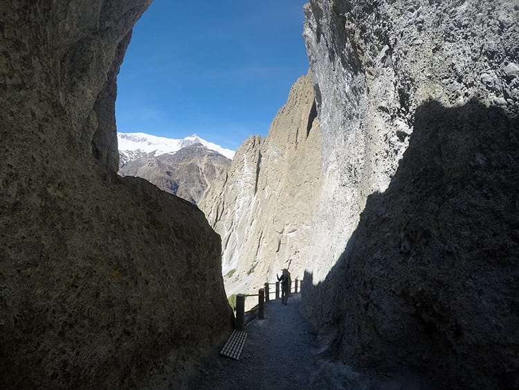 A trekkers walks through a large hole in the boulders where there's a steep path going downhill