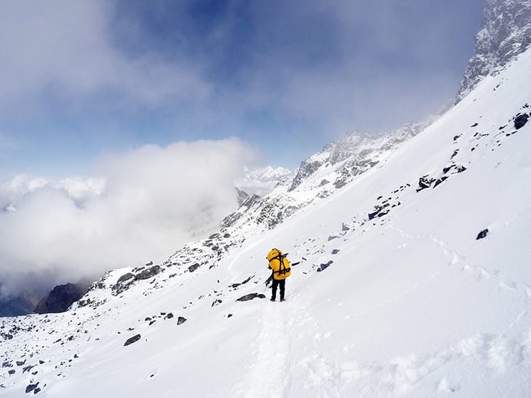 A porter carries a large yellow bag on a small trail covered in snow on the side of a mountain