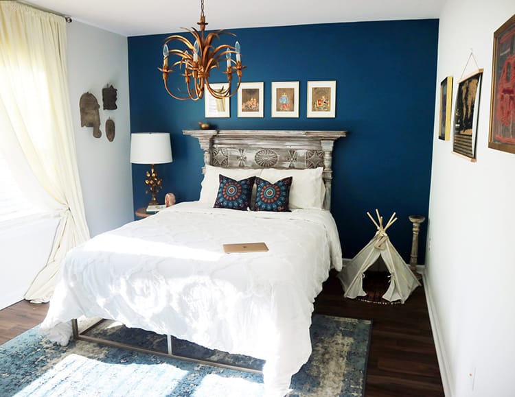Bedroom with one deep turquoise wall, an antique mantle as a headboard, a cream duvet, and souvenirs