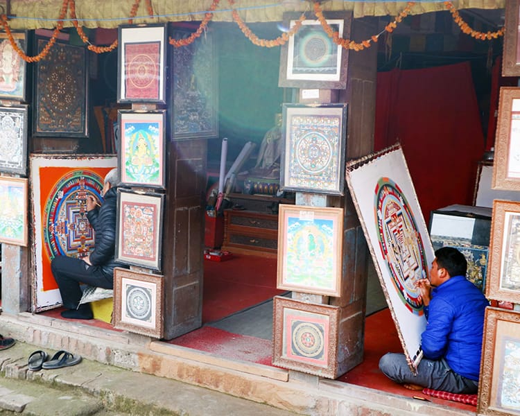 Three people sit in front of a store painting Thangka paintings during the day