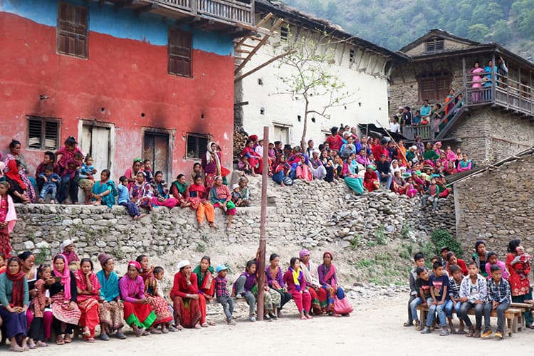 Villagers sit is seats and along walls within the village square wearing brightly colored sarees