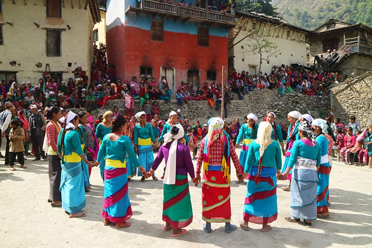 Twenty women hold hands and sing in a circle while wearing brightly colored sarees