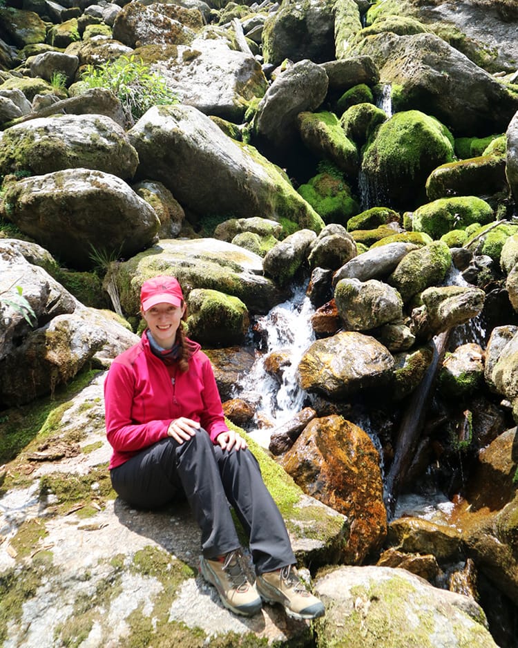 Michelle Della Giovanna from Full Time Explorer sits on some rocks below a waterfall while taking a break from the trek
