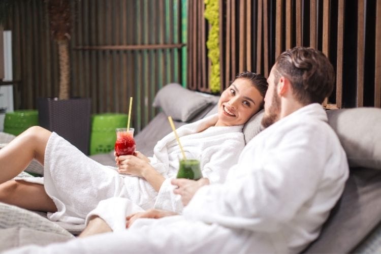 A couple enjoys cocktails while wearing spa robes in lounge chairs - Travel Inspired Stay at Home Date Ideas for Couples
