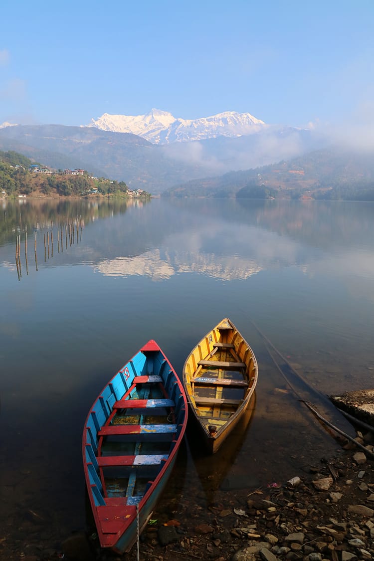 Two colorful boats sit at the edge of Begnas Lake in Pokhara as the Himalaya mountains reflect perfectly in the water