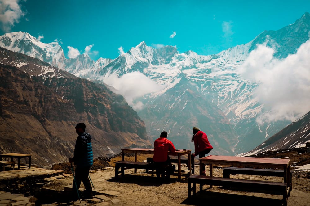 Trekkers sit at a table drinking tea in ABC Base Camp with an incredible view behind them of the fishtail mountain Machhapuchhre